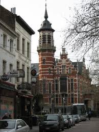 Plombier Jette travaillant près de l'église Notre-Dame de Lourdes, rue commerçante animée bordée de maisons typiques.
